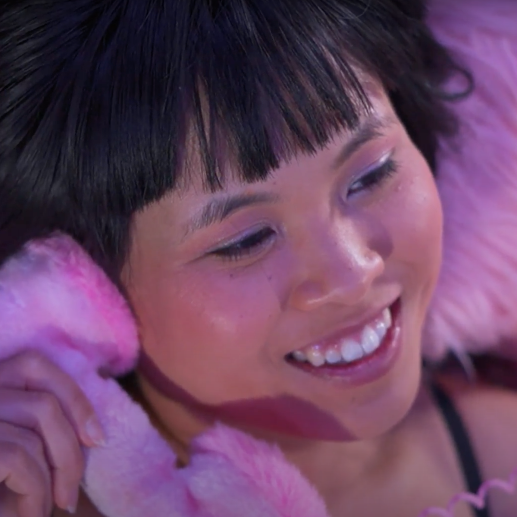 Lena Chen, a Chinese-American woman with bangs and perfect eyebrows, smiling off into the distance while chatting on a pink furry phone. The image is flirty.