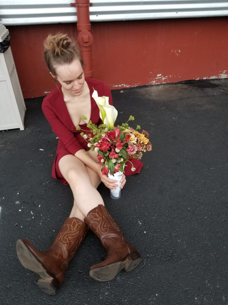 Maggie Oates, a white femme, sits in a parking lot with cowboy boots and a bouquet of flowers